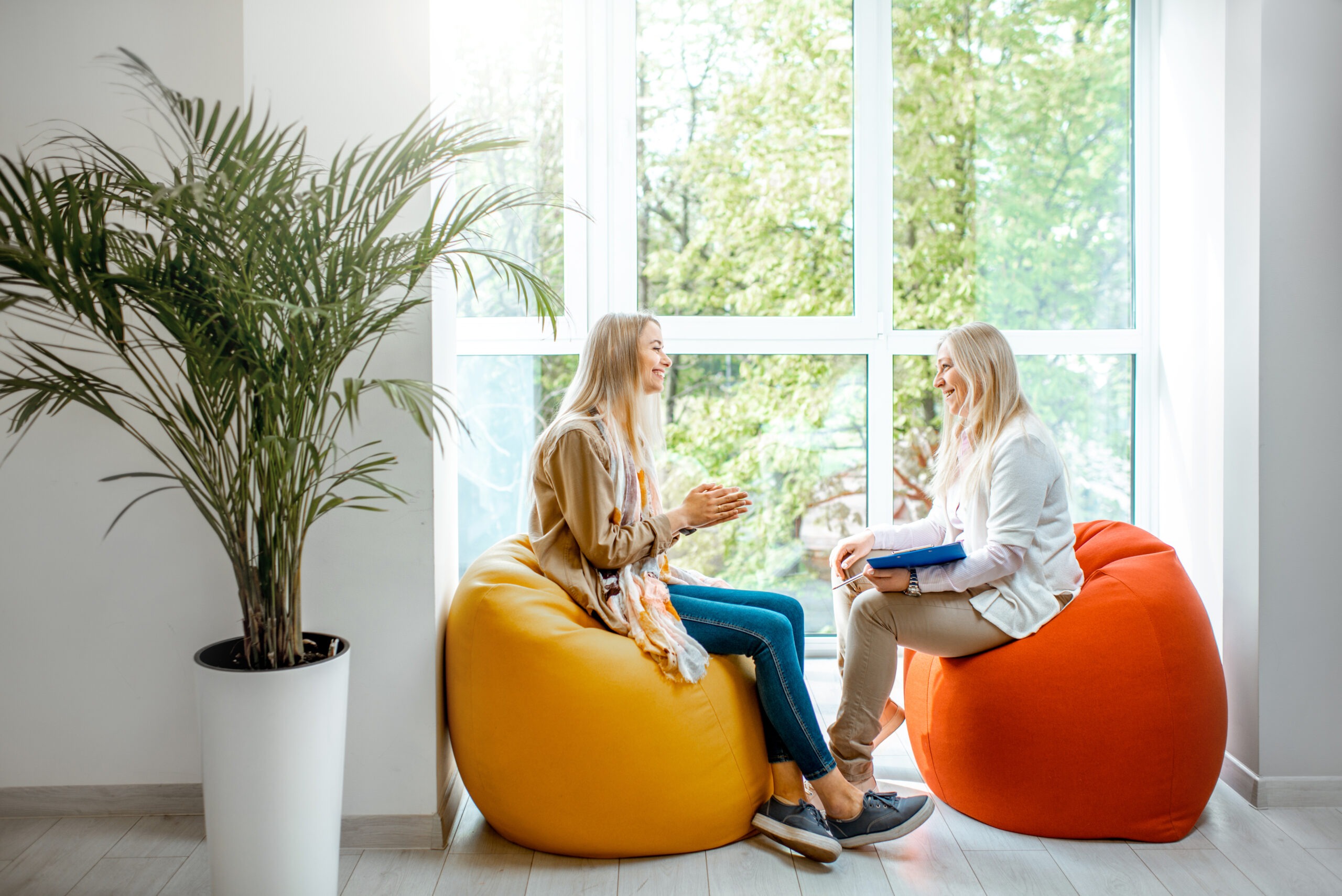 Two women engage in a lively conversation while seated on colorful bean bags by a large window overlooking greenery.